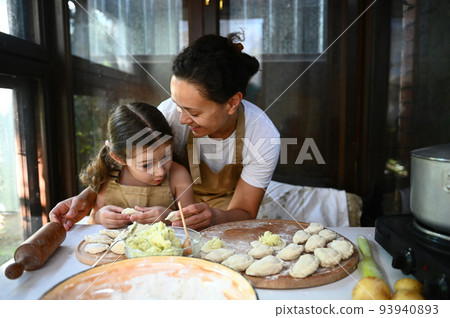 Loving mother hugging her adorable daughter, standing together at a table with dumplings, enjoying cooking varenyky Loving mother hugging her adorable daughter, standing together at a table with dumplings, enjoying cooking varenyky 93940893