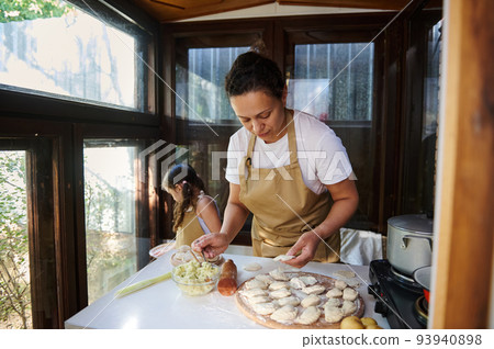 Multiethnic woman, young loving mother in beige chef's apron, preparing dumpling or pelmeni dough or Ukrainian varenyky 93940898