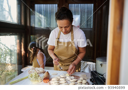 Beautiful housewife puts mashed potatoes on a rolled out dough, enjoys cooking dumplings for lunch with her daughter. 93940899