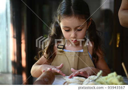 Close-up charming child girl with two ponytails, in beige chef's apron, molding dumplings, cooking vareniki with her mom 93943567