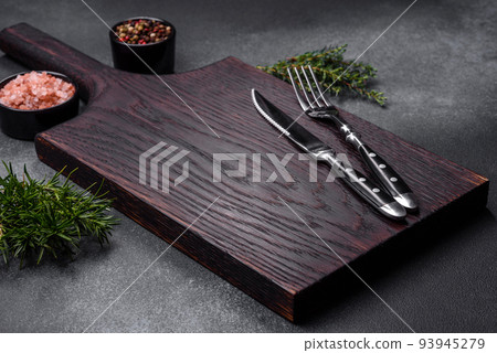Wooden cutting board, salt, pepper, spices and herbs on a dark concrete background 93945279