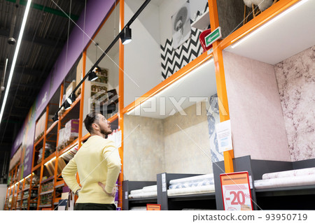 a man chooses finishing materials for repairs in his house next to the racks in a construction a man chooses finishing materials for repairs in his house next to the racks in a construction 93950719