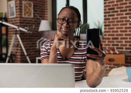 Young african american woman taking selfie on smartphone, smiling at front camera. Freelancer making portrait photo on mobile phone at home office workplace, remote worker talking at video call Young african american woman taking selfie on smartphone, smiling at front camera. Freelancer making portrait photo on mobile phone at home office workplace, remote worker talking at video call 93952529