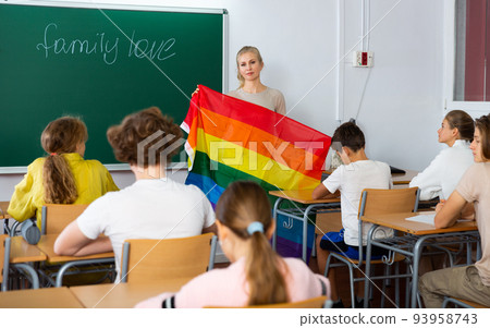 Progressive female teacher discussing with teenage students about LGBT social movements in classroom, holding rainbow flag Progressive female teacher discussing with teenage students about LGBT social movements in classroom, holding rainbow flag 93958743