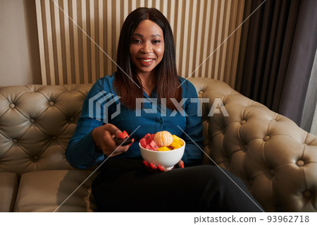 Smiling Black woman sitting on couch with bowl of yeah fruits and turning on tv to watch new show Smiling Black woman sitting on couch with bowl of yeah fruits and turning on tv to watch new show 93962718