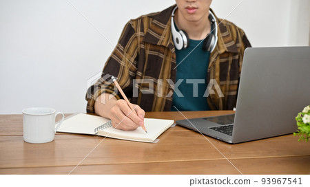 A hipster young Asian male college student using laptop and taking notes on his book. cropped 93967541