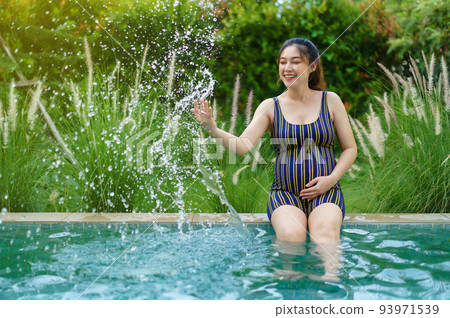 cheerful pregnant woman in swimsuit sitting on edge of swimming pool while playing water splashing 93971539