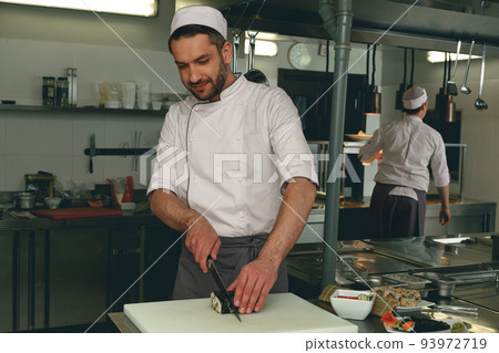 Smiling Male Chef in uniforms preparing sushi in a kitchen of asian restaurant Smiling Male Chef in uniforms preparing sushi in a kitchen of asian restaurant 93972719