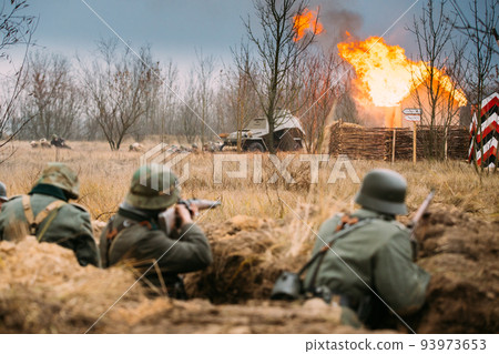 Re-enactors Armed Rifles And Dressed As World War Ii German Wehrmacht Infantry Soldiers Fighting Defensively In Trench. Defensive Position. Fight Against Combat Vehicle. Building On Fire On Background 93973653