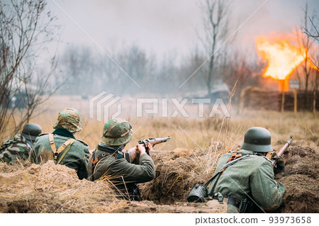 Re-enactors Armed Rifles And Dressed As World War Ii German Wehrmacht Infantry Soldiers Fighting Defensively In Trench. Defensive Position. Smokescreen. Building On Fire On Background 93973658