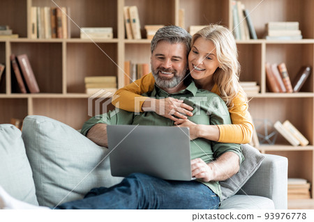 Smiling Middle Aged Spouses Spending Time With Laptop In Living Room Smiling Middle Aged Spouses Spending Time With Laptop In Living Room 93976778