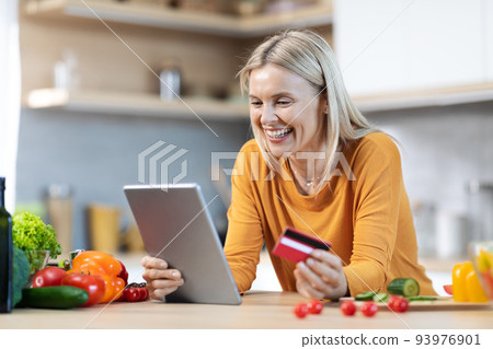Happy blonde woman buying food online, using digital pad Happy blonde woman buying food online, using digital pad 93976901