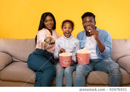 Emotional african american family of three watching movie, young parents sitting with daughter on couch, eating popcorn Emotional african american family of three watching movie, young parents sitting with daughter on couch, eating popcorn 93976931