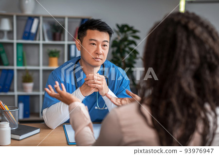 Serious millennial chinese guy doctor in uniform listens symptoms of african american lady patient in office 93976947