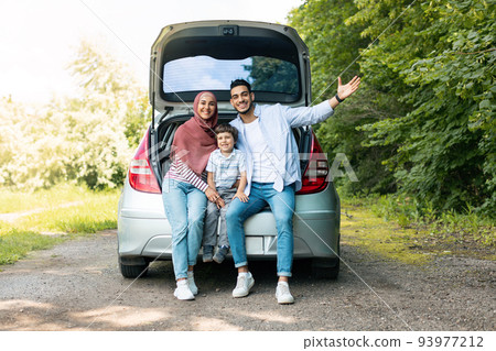 Positive auto journey. Happy millennial arabic guy and lady in hijab with baby, sit in car trunk and gesturing Positive auto journey. Happy millennial arabic guy and lady in hijab with baby, sit in car trunk and gesturing 93977212