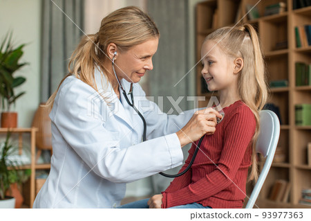Happy little girl having checkup at pediatrician, child smiling while doctor checking up her chest with stethoscope Happy little girl having checkup at pediatrician, child smiling while doctor checking up her chest with stethoscope 93977363