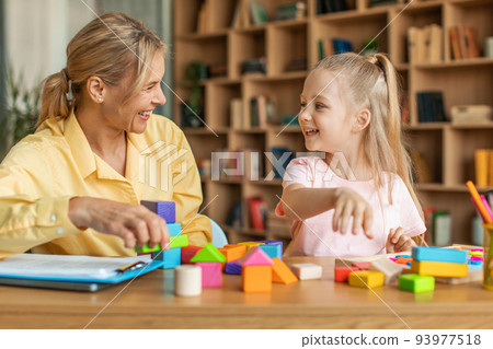 Positive kids psychology. Professional female therapist with notepad watching little girl playing with colorful blocks Positive kids psychology. Professional female therapist with notepad watching little girl playing with colorful blocks 93977518