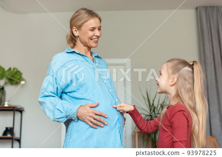 Happy little girl touching pregnant mother's belly, spending time at home together, looking at each other and smiling 93978225