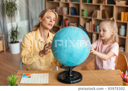 Professional preschool teacher teaching cute girl about globe, having private home class, sitting at desk in living room 93978302