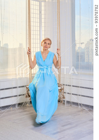 Middle age woman chilling at home sitting on hanging swing in long blue dress Middle age woman chilling at home sitting on hanging swing in long blue dress 93978782