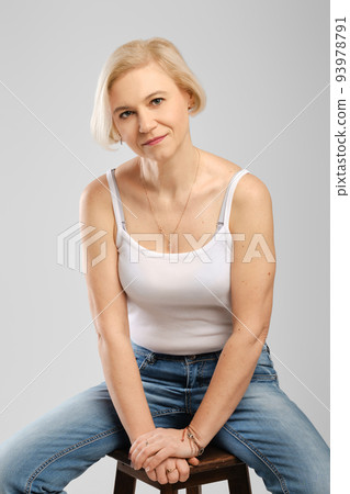 Barefoot senior woman in jeans and tank top sitting on tall chair in studio Barefoot senior woman in jeans and tank top sitting on tall chair in studio 93978791