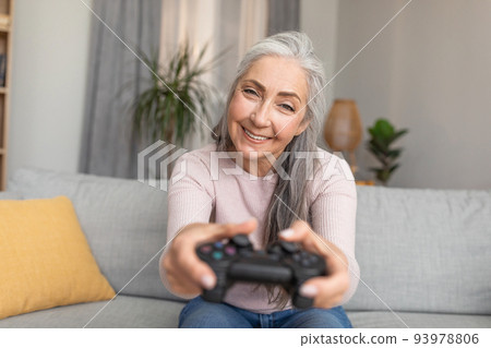Glad smiling caucasian old gray-haired female with joystick playing online game in living room interior 93978806