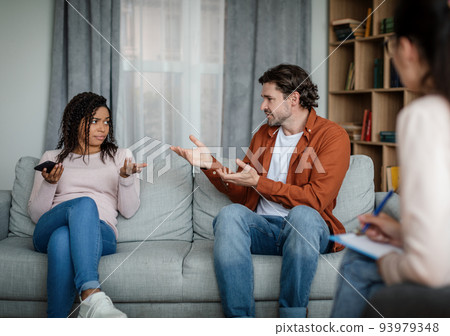 Unhappy european husband swears at black wife with smartphone on consultation with psychologist in clinic Unhappy european husband swears at black wife with smartphone on consultation with psychologist in clinic 93979348