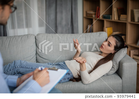 Sad european millennial lady patient lies on couch and talks on consultation, male doctor makes notes 93979441