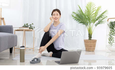 Beautiful Asian woman watching yoga teacher and light exercises in her living room at home, Watch live teaching or VDO on laptop, Dumbbells and a protein shake or bottle of water, Healthy exercise. 93979605