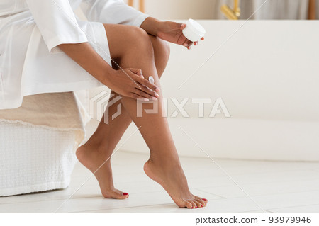 Unrecognizable black female in white silk robe applying moisturising cream on legs 93979946