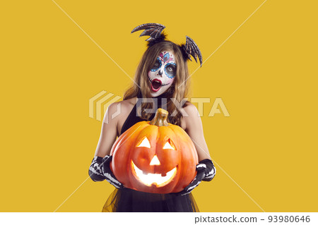 Girl in Halloween costume holding Jack o lantern and looking at camera with surprised expression Girl in Halloween costume holding Jack o lantern and looking at camera with surprised expression 93980646