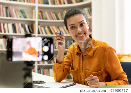 Excited mixed race female tutor having online lecture, talking to smartphone camera, making video call in library Excited mixed race female tutor having online lecture, talking to smartphone camera, making video call in library 93980717