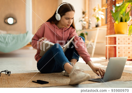 Concentrated teenage girl reading book in front of laptop, getting ready for exam, sitting on floor at home 93980970