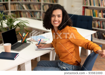Working on research project. Happy female student using laptop at library, writing in notepad, smiling at camera 93981000