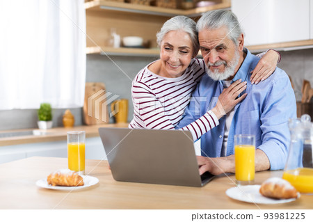 Smiling senior couple using laptop in kitchen while having breakfast together 93981225