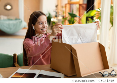 Happy customer. Happy teen girl opening and unpacking parcel cardboard box, sitting at table at home and smiling 93981256
