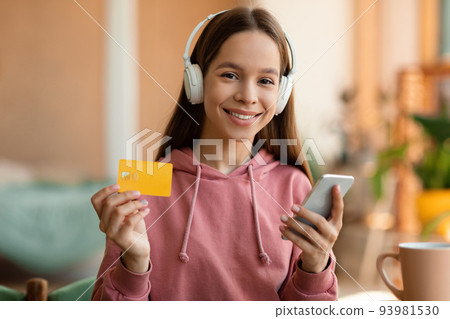 Happy teen girl holding credit card and paying with smartphone, listening music in wireless headphones, sitting at home Happy teen girl holding credit card and paying with smartphone, listening music in wireless headphones, sitting at home 93981530