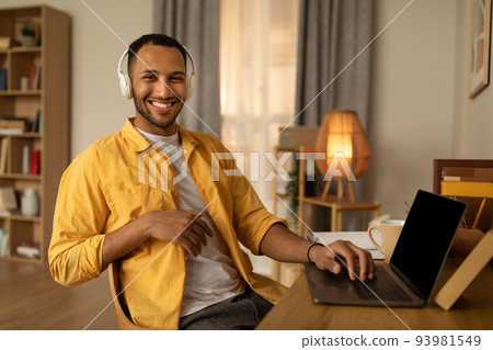 Young happy black man in headphones using laptop pc with mockup, working online at home, copy space Young happy black man in headphones using laptop pc with mockup, working online at home, copy space 93981549