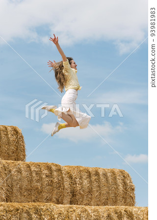 Young woman jumps on the haystack with her hands raised high. Countryside holiday on the farm, blue sky. Hay harvesting. 93981983