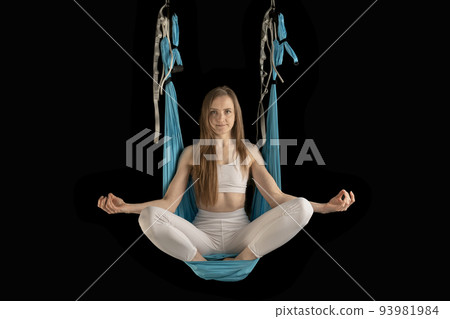 Athletic girl meditates sitting in sports hammock. Fly yoga. Portrait of young woman during meditation on black background. Athletic girl meditates sitting in sports hammock. Fly yoga. Portrait of young woman during meditation on black background. 93981984