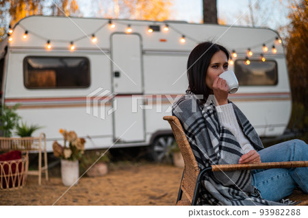 Caucasian woman in a knitted hat wrapped in a plaid and drinks a warming drink outdoors. Travel in a motor home in the fall. 93982288