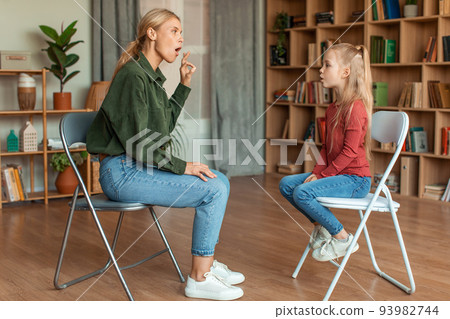 Preparing for school. Pretty girl studying with professional woman speech therapist, pronouncing sound O at classroom Preparing for school. Pretty girl studying with professional woman speech therapist, pronouncing sound O at classroom 93982744