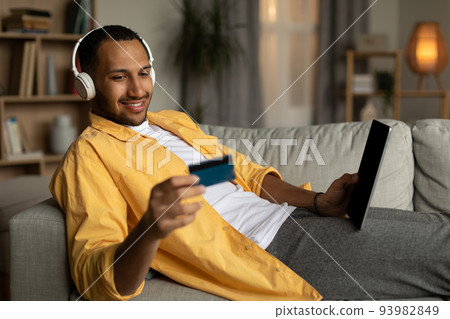 Young black man in headphones buying goods online, using tablet and credit card, enjoying shopping on web at home Young black man in headphones buying goods online, using tablet and credit card, enjoying shopping on web at home 93982849