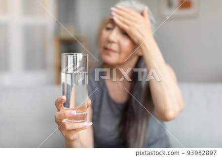 Sad caucasian elderly gray-haired woman presses her hand to forehead and holds glass of water, suffers from headache Sad caucasian elderly gray-haired woman presses her hand to forehead and holds glass of water, suffers from headache 93982897