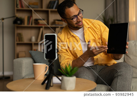 Cheerful young black male blogger recording video review of new digital tablet on cellphone at home, mockup 93982945