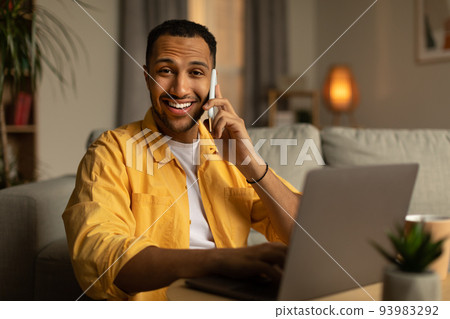 Handsome young black man in casual wear talking on smartphone, enjoying conversation at home office Handsome young black man in casual wear talking on smartphone, enjoying conversation at home office 93983292