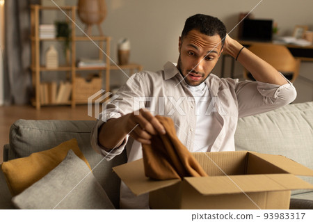 Dissatisfied buyer. Frustrated black man unboxing package after delivery, holding clothes, sitting on couch at home 93983317