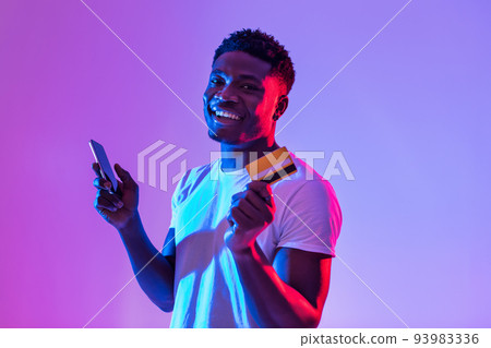 Young black man holding cellphone and credit card, using banking or shopping mobile app in neon light 93983336