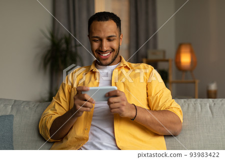 Happy young black man using smartphone, chatting online, sitting on couch at home Happy young black man using smartphone, chatting online, sitting on couch at home 93983422