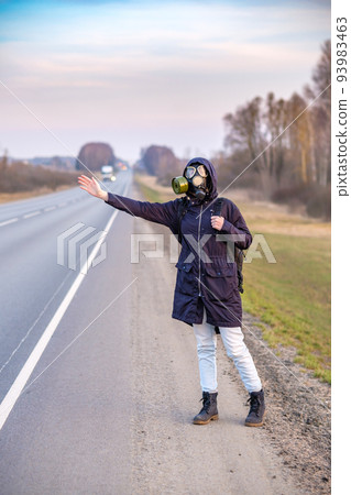 A girl in dark clothes, a gas mask and a hood on her head raises her hand and tries to stop a passing car. 93983463